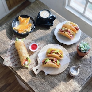 May include: A miniature food display featuring a rolled wrap, two tacos on a wooden board, a plate of two tacos, a bowl of yellow chips, a black mug with a white swirl, a small red sauce container, a white condiment jar, and a small succulent.