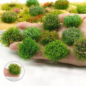 Pode incluir: Um conjunto de tufos esféricos de relva artificial em tons de verde, com pequenos detalhes florais coloridos. Alguns são segurados na mão, enquanto outros estão dispostos em uma paisagem em miniatura, ideais para modelismo ou projetos de artesanato.