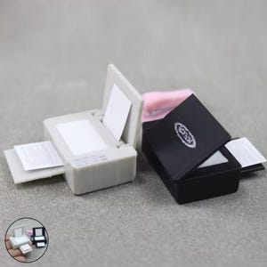 Puede incluir: Mini impresoras en gris y negro. La impresora gris tiene una hoja de papel que sale por delante y otra dentro. La impresora negra tiene un diseño blanco en la parte superior y una hoja de papel que sale por delante.