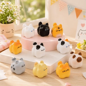 Può includere: Una collezione di dieci piccole statuette di animali in ceramica dalle forme arrotondate, in vari colori tra cui bianco, nero, arancione, giallo e grigio. Ogni statuetta presenta un volto semplice con due punti per gli occhi e una piccola bocca, disposte su ripiani.