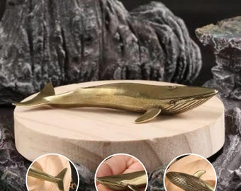 May include: A brass whale figurine with detailed fins and tail, resting on a wooden platform. The whale's body has etched lines, and the overall design is a polished gold color. The figurine is a decorative item.
