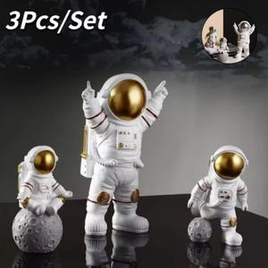 Puede incluir: Conjunto de tres figuras de astronautas blancas con cascos dorados. Un astronauta está sentado en una luna gris, otro está de pie con los brazos levantados y el tercero está arrodillado con una luna gris. El texto "3Pcs/Set" está en la parte superior.
