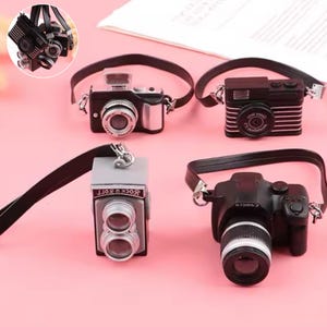 May include: A collection of miniature cameras in black and silver, each with a black strap. The designs vary, including a retro model with horizontal lines and a modern DSLR style. The background is a pale pink.