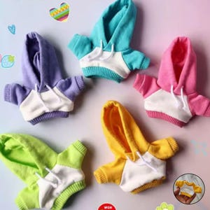 Op de afbeelding: Een verzameling miniatuurhoodies in verschillende kleuren, waaronder paars, blauwgroen, roze, geel en groen. Elke hoodie heeft een witte torso, een capuchon met trekkoord en bijpassende mouwen. De hoodies zijn ontworpen voor poppen of klein speelgoed.