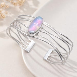 May include: A silver cuff bracelet with an oval, iridescent gemstone. The bracelet has a multi-strand design with a central gemstone setting. The gemstone displays pink, blue, and white hues. The bracelet is a stylish piece of jewellery.