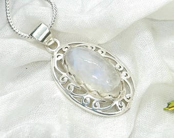 May include: A silver necklace with an oval pendant featuring a large, iridescent moonstone. The pendant is set in an ornate, openwork frame. The necklace rests on a white, textured fabric, with a yellow flower in the background.