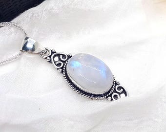 May include: A silver pendant necklace featuring an oval, iridescent moonstone. The moonstone is set in a decorative, filigree-style frame. The necklace rests on a white fabric surface, with a black box in the background.