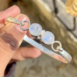May include: A silver bangle bracelet with two round moonstone cabochons. The bracelet has a simple design with a hinged clasp. The moonstones have a milky appearance. The bracelet's diameter is approximately 6 cm.