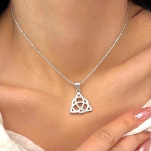 May include: A silver necklace featuring a Celtic knot pendant. The pendant has a triangular shape with an interwoven design. The necklace is worn by a person wearing a white shirt.