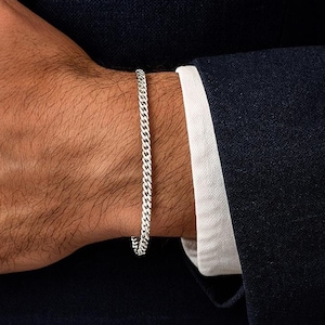 May include: A silver curb chain bracelet. The bracelet is worn on a wrist, with a dark blue suit jacket and white shirt cuff in the background. A classic, understated piece of jewellery.