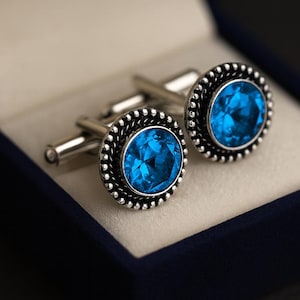 Puede incluir: Un par de gemelos plateados con grandes piedras preciosas ovaladas de color azul brillante. Las piedras están engastadas en un marco plateado decorativo con cuentas. Presentados en una caja de terciopelo azul oscuro.