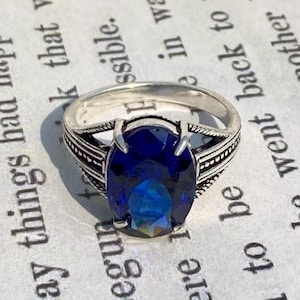 Op de afbeelding: Een zilveren ring met een ovale, diepblauwe edelsteen. De ring heeft een sierlijk ontwerp met gedetailleerde patronen op de band. De edelsteen heeft een meerkleurig uiterlijk, dat verschillende blauwtinten weerspiegelt. De ring staat tegen een achtergrond van gedrukte tekst.