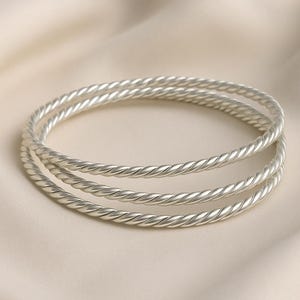 Può includere: Tre braccialetti color argento con un design a corda intrecciata. I braccialetti sono impilati l'uno sull'altro, creando un effetto a strati. Lo sfondo è di un colore morbido e neutro.