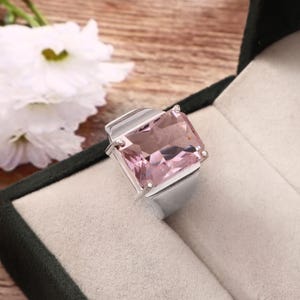 May include: A silver ring with a large, rectangular, light pink gemstone. The ring is set in a simple, modern style, with a thick band and a bezel setting. The gemstone is a soft, pastel pink color. The ring is displayed in a black jewelry box.
