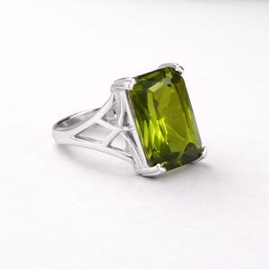 Op de afbeelding: Een zilveren ring met een grote, rechthoekige, lichtgroene edelsteen. De ring heeft een geometrisch ontwerp aan de zijkanten en de edelsteen is gezet in een zilveren band. De ring wordt getoond tegen een witte achtergrond.