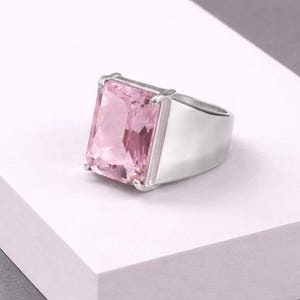 May include: A silver ring with a large, rectangular pink gemstone. The ring has a thick band and a modern, minimalist design. The gemstone is a light pink color and is cut with multiple facets.