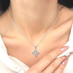 Op de afbeelding: Een zilverkleurige ketting met een Keltische knoop hanger. De hanger heeft een vierpuntig ontwerp met ingewikkeld openwerk. De ketting wordt gedragen en de ketting is fijn en delicaat.
