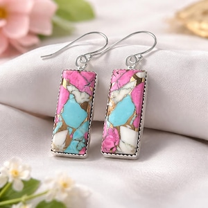 May include: A pair of rectangular earrings with a silver-coloured metal frame. The earrings feature a mosaic design with pink, turquoise, white, and gold-coloured stone inlays. The earrings have a hook closure.