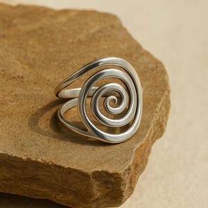 Peut inclure: Une bague en argent avec un motif en spirale. La bague présente un motif tourbillonnant au centre, créant un look unique et accrocheur. La bague est en métal brillant et est posée sur un fond neutre.