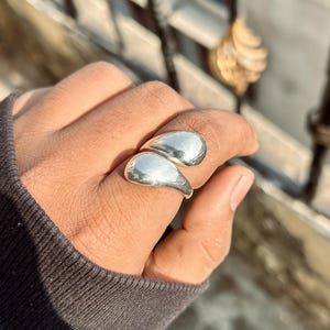 May include: A silver ring with a unique design featuring two teardrop-shaped elements. The ring is worn on a hand, showcasing its polished, reflective surface. The ring's design is modern and eye-catching, suitable for everyday wear.