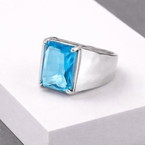 Op de afbeelding: Een zilveren ring met een grote, rechthoekige, lichtblauwe edelsteen. De ring heeft een gepolijst, modern design met een brede band. De edelsteen is gezet in een bezel setting, die de levendige kleur en helderheid benadrukt.