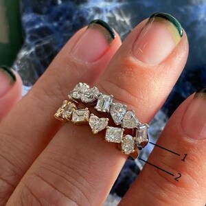 May include: Two gold rings with various-shaped clear gemstones. The rings are stacked on a finger, showcasing the different cuts and sizes of the stones. The gemstones include square, heart, and rectangular shapes.