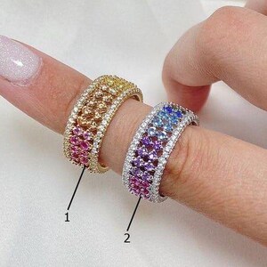 May include: Two rings with colorful gemstones. One ring is gold-toned with pink, yellow, and red stones. The other ring is silver-toned with blue, purple, and pink stones. Both rings have a double row of stones.