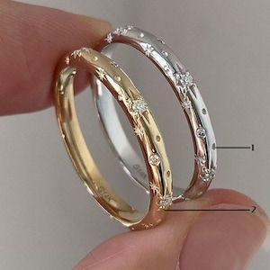 May include: Two rings, one gold and one silver, are held between fingers. The gold ring is engraved with stars and small diamonds. The silver ring has a similar design. Both rings have a delicate, celestial theme.