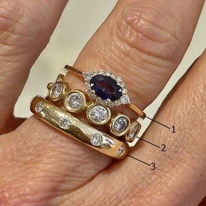 Puede incluir: Una mano con tres anillos de oro. El anillo superior tiene una piedra preciosa azul en forma de marquesa rodeada de pequeños diamantes. El anillo central tiene diamantes engarzados. El anillo inferior es una banda gruesa con diamantes engarzados espaciados.