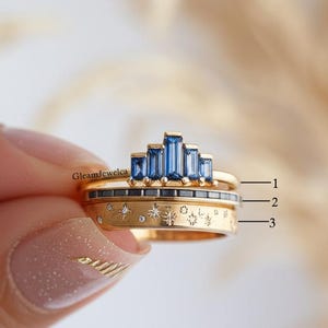 May include: Three gold rings with blue and clear gemstones. The top ring features a row of blue baguette-cut stones. The middle ring has a row of small black stones. The bottom ring has a celestial design with stars and small diamonds.