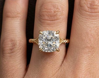 May include: A gold ring featuring a large, cushion-cut clear gemstone held by four prongs. The band has a twisted rope texture on the sides and intricate metal detailing where the band meets the central stone setting, creating a refined appearance.