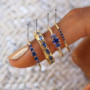 Könnte beinhalten: Vier goldene Ringe mit blauen Edelsteinen. Die Ringe weisen verschiedene Designs auf, darunter ein Band mit ovalen Steinen, ein Band mit einer Reihe kleiner Steine und ein Band mit markisenförmigen Steinen. Die Ringe befinden sich auf einem Finger.