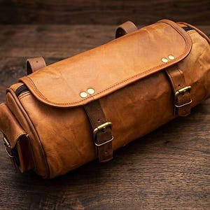 Può includere: Una borsa cilindrica in pelle color cuoio con una grande patta frontale fissata da due cinghie regolabili con fibbie e rivetti in metallo color ottone. La borsa presenta una tasca laterale e una chiusura a zip con una finitura liscia e testurizzata.