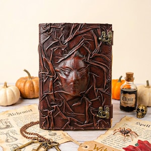 May include: A dark brown leather journal with a raised face design and metal clasp. The journal is labelled "Witch's Grimoire" and is surrounded by Halloween-themed items, including pumpkins, keys, and a small bottle labelled "Elixir".