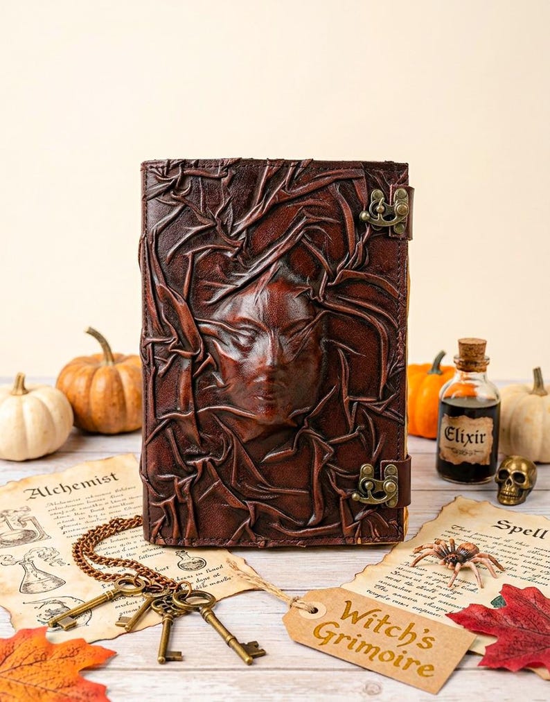 May include: A dark brown leather journal with a raised face design and metal clasp. The journal is labelled "Witch's Grimoire" and is surrounded by Halloween-themed items, including pumpkins, keys, and a small bottle labelled "Elixir".