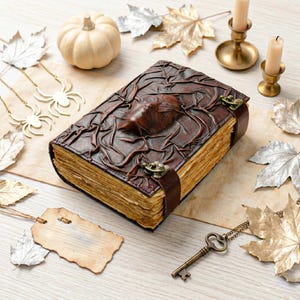 May include: A brown leather journal with an embossed design, secured by a strap and clasp. The aged pages and decorative elements like a pumpkin, spider pendants, and candles suggest a fall or Halloween theme.