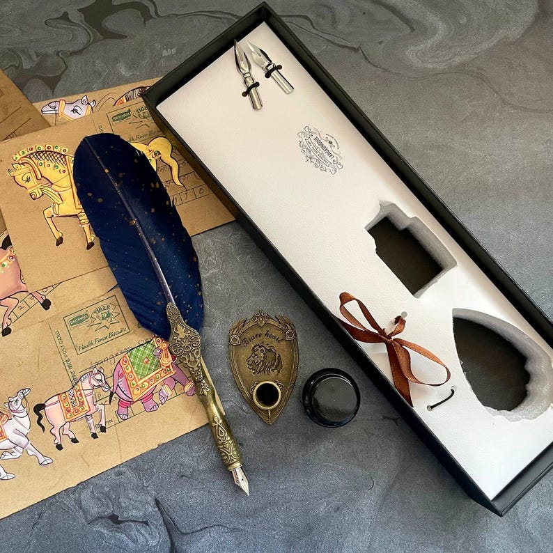 May include: A vintage calligraphy set featuring a blue feather pen, inkwell, and nibs, presented in a black box. The set includes a brass pen holder and is accompanied by decorative paper with illustrations of horses.