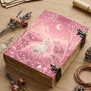 Può includere: Un diario con copertina rigida caratterizzato da un'illustrazione rosa di un unicorno bianco in una foresta con funghi, fiori e una luna crescente. Il libro è fissato con due fermagli in metallo scuro e contiene pagine di carta color crema invecchiata.