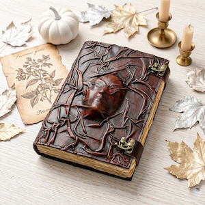 May include: A brown leather journal with a raised face design and metal clasp. The journal is surrounded by decorative elements, including a small white pumpkin, candles, and autumn leaves. A vintage botanical print is also present.