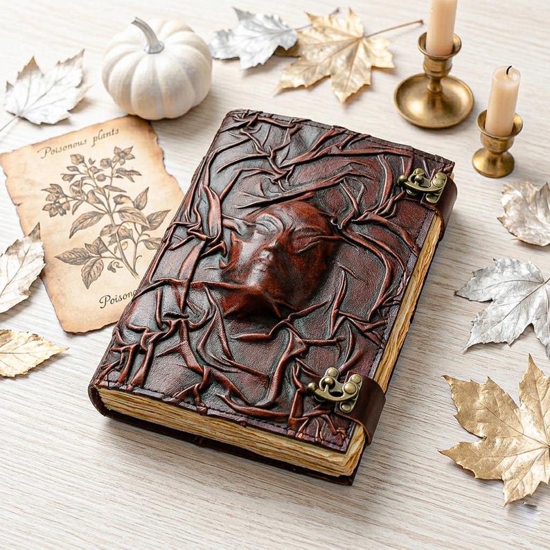 May include: A brown leather journal with a raised face design and metal clasp. The journal is surrounded by decorative elements, including a small white pumpkin, candles, and autumn leaves. A vintage botanical print is also present.