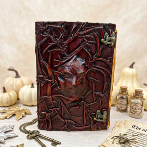 May include: A dark brown leather journal with an embossed face design. It features a metal clasp and is displayed with antique keys, potion bottles, and aged paper. The theme is gothic and fantasy, with a vintage aesthetic.