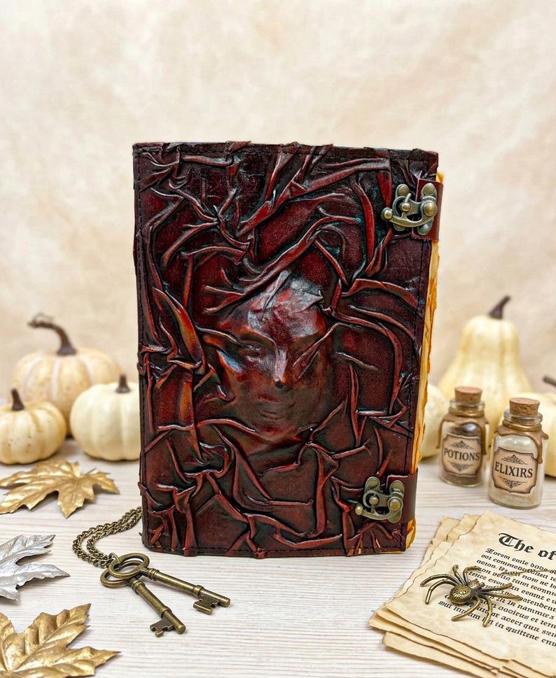 May include: A dark brown leather journal with an embossed face design. It features a metal clasp and is displayed with antique keys, potion bottles, and aged paper. The theme is gothic and fantasy, with a vintage aesthetic.