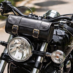 May include: Close-up of a black motorcycle with a leather storage bag mounted on the handlebars. The motorcycle has chrome accents, including the headlight casings and handlebars. The bag is secured with gold-colored buckles.