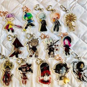 May include: Collection of acrylic keychains featuring various cartoon characters. Each keychain has a unique design with vibrant colors and attached to a metal keyring with a star-shaped charm. The keychains are displayed on a white, quilted surface.