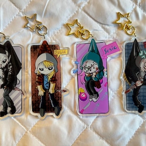 May include: Four acrylic keychains featuring cartoon characters with unique designs. Each keychain has a gold star-shaped charm and a metal clasp. The characters are depicted with various outfits and accessories, including knives and name tags.