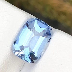 May include: A light blue gemstone with a rectangular cushion cut. The gem has multiple facets that reflect light, creating a sparkling effect. The stone is set against a white background.