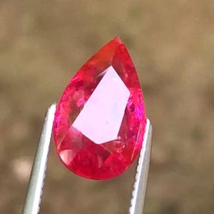 Puede incluir: Una gema roja facetada en forma de pera, sujeta con pinzas plateadas. La gema tiene una superficie brillante y reflectante con una gama de tonos rojos. La gema es una pieza vibrante y llamativa.