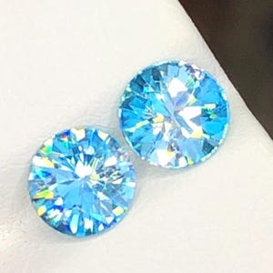 May include: Two round, faceted, light blue gemstones. The gems have a brilliant cut, reflecting light with a rainbow of colors. The stones are set against a white background, highlighting their clarity and sparkle.