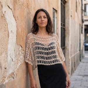 Peut inclure: Un haut en crochet beige présentant un motif à mailles ouvertes avec de larges bandes horizontales et des manches courtes, porté sur une robe noire sans manches. Le haut possède une large encolure arrondie et une coupe ample et carrée.