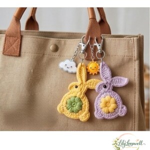 May include: Two crochet bunny keychains attached to a tan canvas tote bag. One bunny is yellow with a green flower center, and the other is light purple with a yellow flower center. Each features a small metal charm of a smiling cloud or sun.